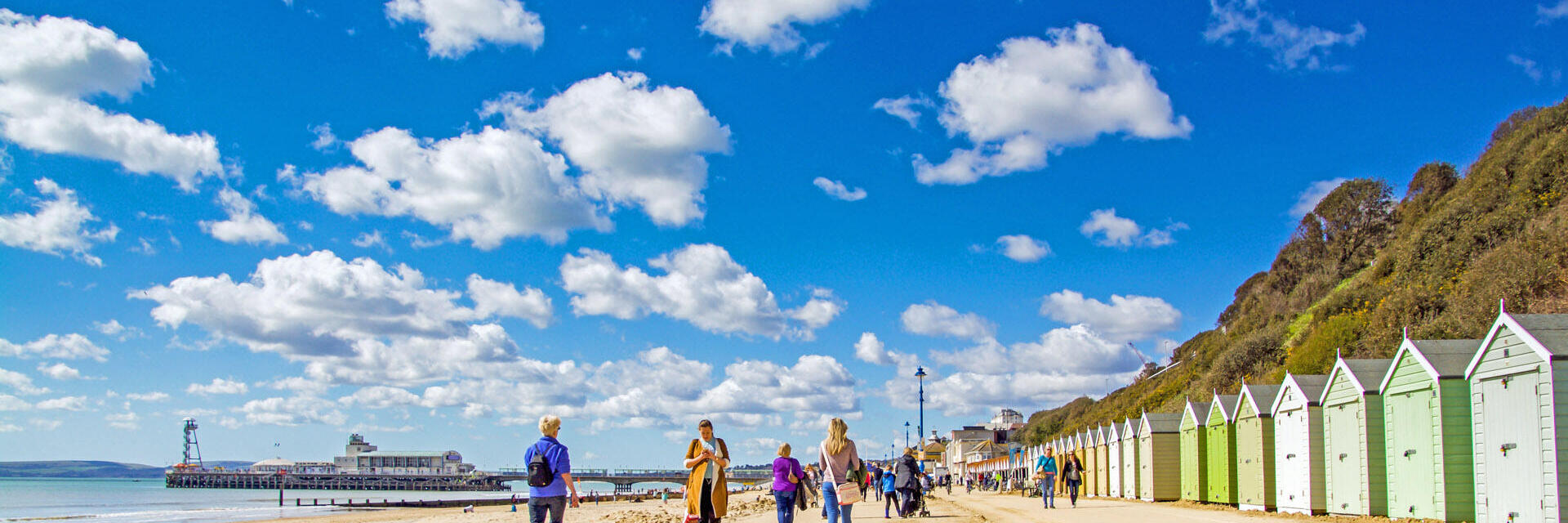 People walking along the coast next to colourful beach huts on a sunny day.