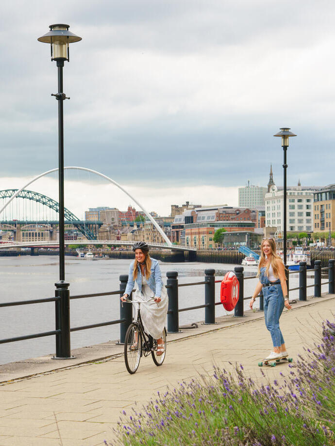 Two people cycling and skateboarding on a path along the River Tyne in Newcastle.