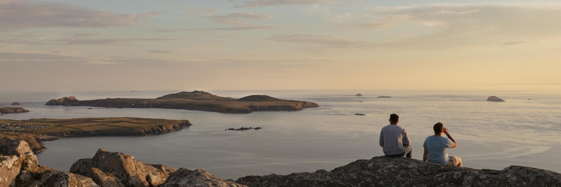 Zwei Männer sitzen auf einer Felsklippe und blicken in der Abenddämmerung auf das Meer