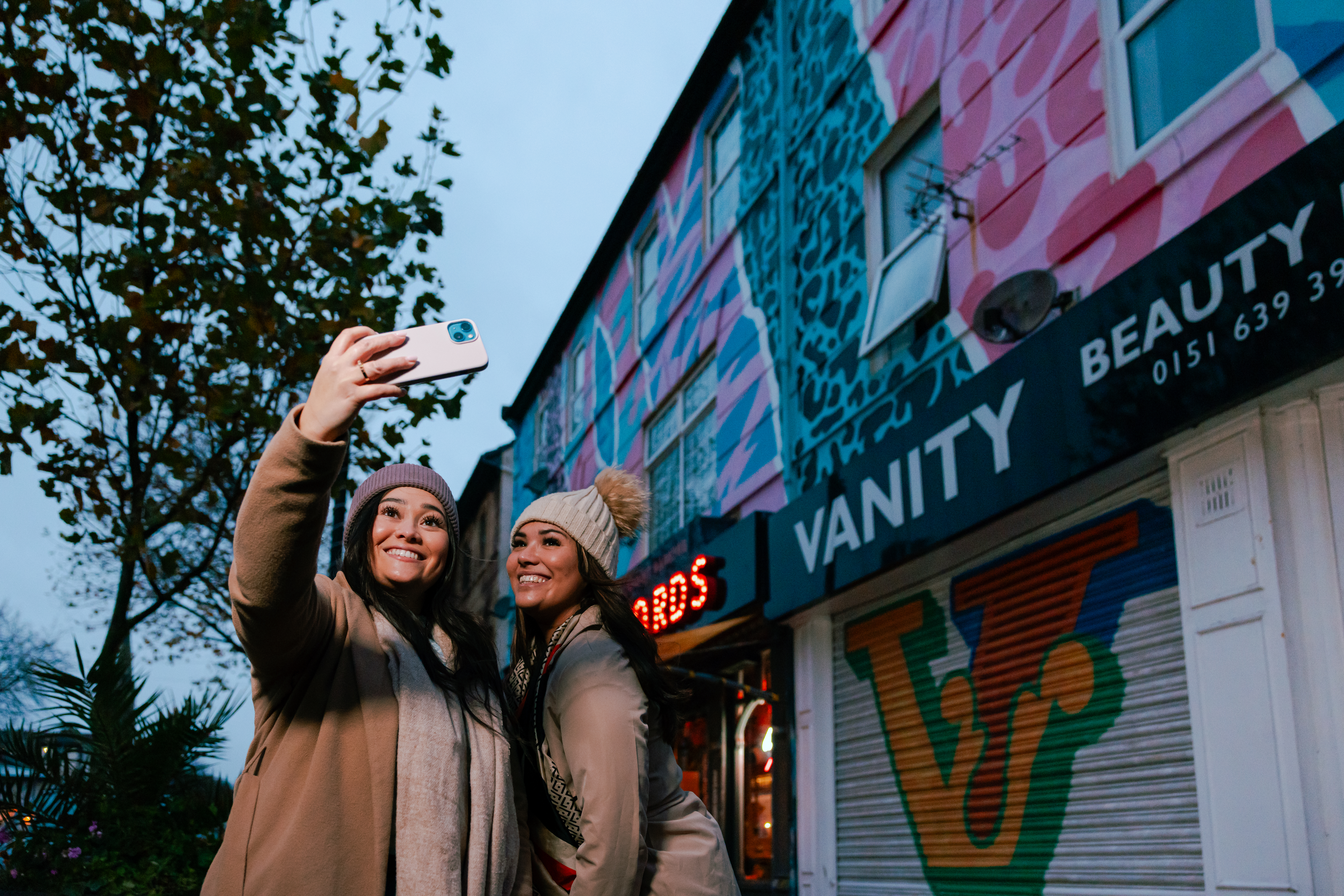 Two women pose for a selfie in front of a row of shops