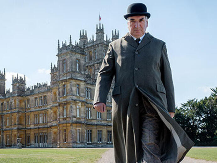 A person in period clothing walks in front of a grand historic mansion with ornate architecture and landscaped grounds.