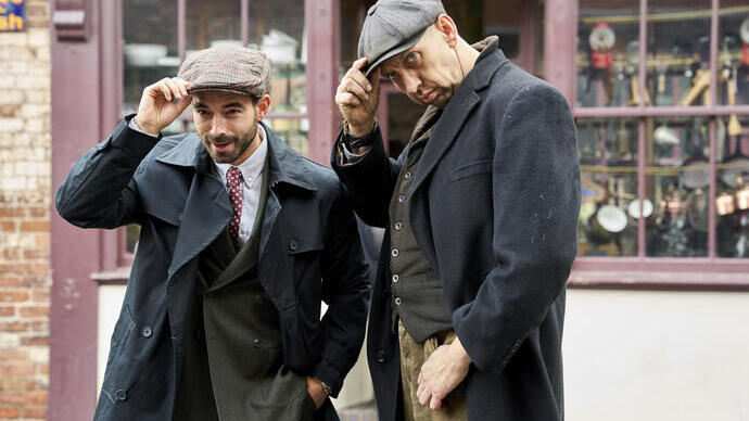 Two men in vintage clothing and flat caps pose outside a shop with a display window and brick wall backdrop.