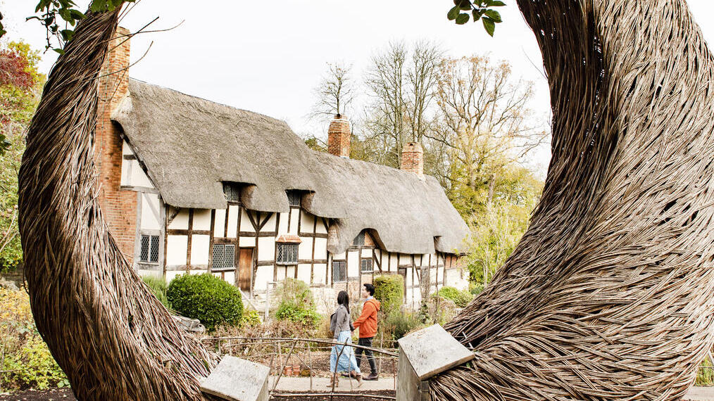 Couple walking near a cottage seen through a large willow crescent shaped sculpture in a garden