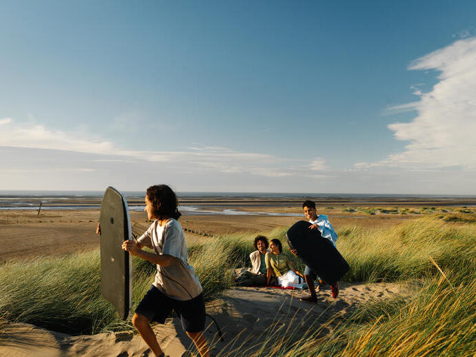 A family enjoys a day at the beach, with children carrying bodyboards and adults relaxing on sand dunes under a blue sky.