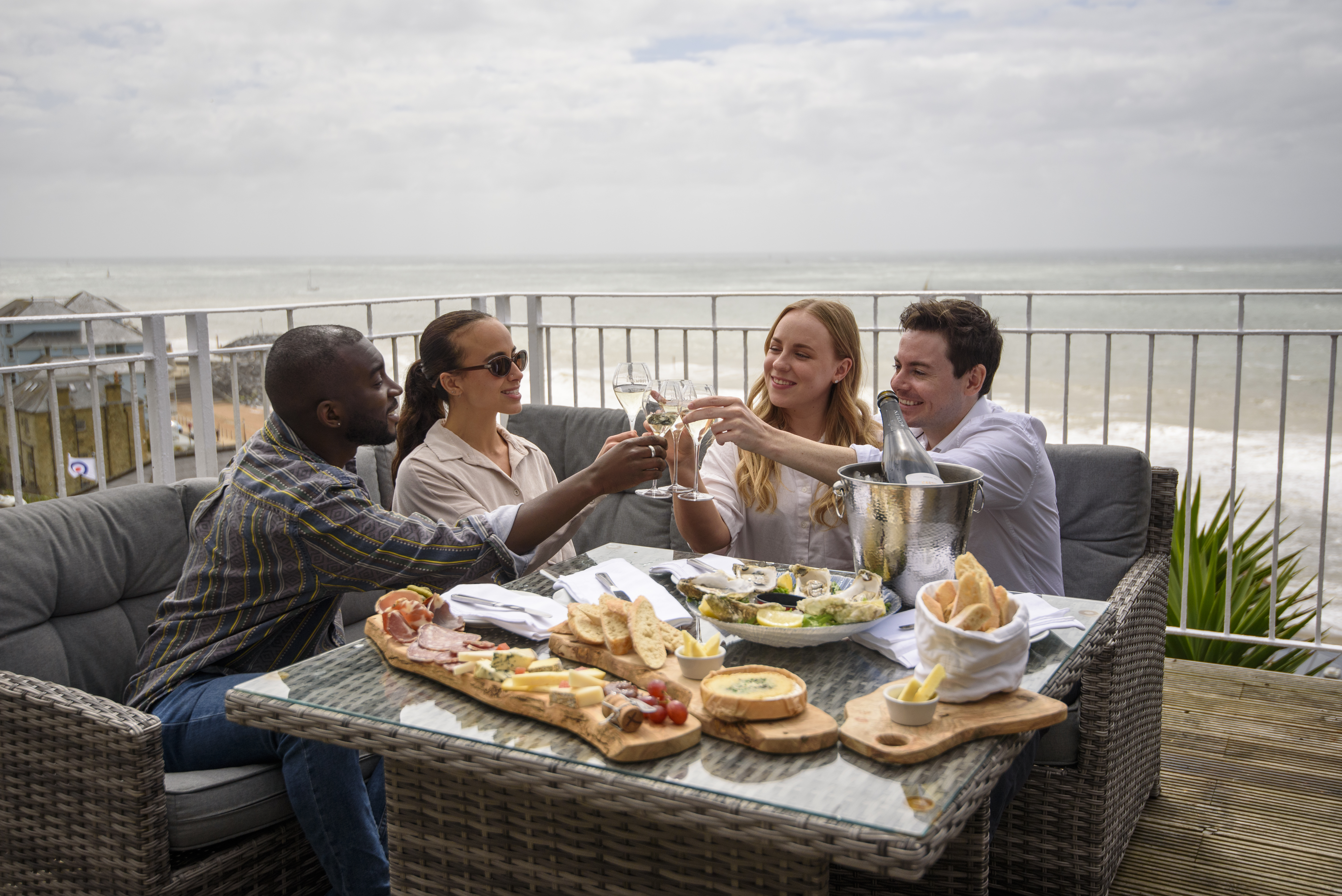 Two men and two women laugh together while sat on a hotel balcony with the sea beyond