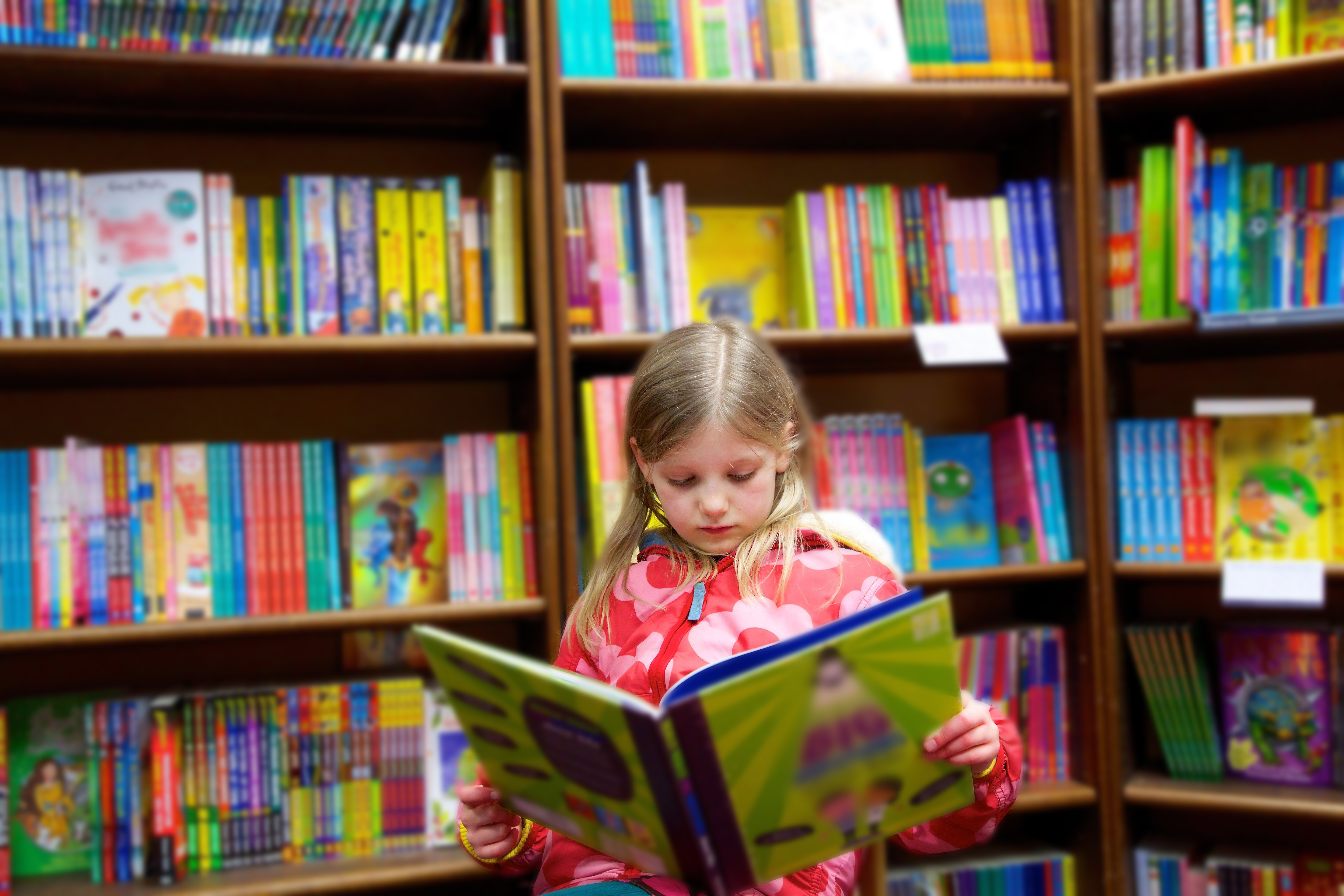 Young girl reading a book in a colorful library with many children's books on shelves in the background.