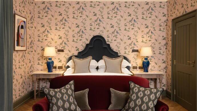 Chambre d'hôtel luxueuse avec un canapé en velours rouge au pied d'un lit et un papier peint à fleurs sur les murs derrière.