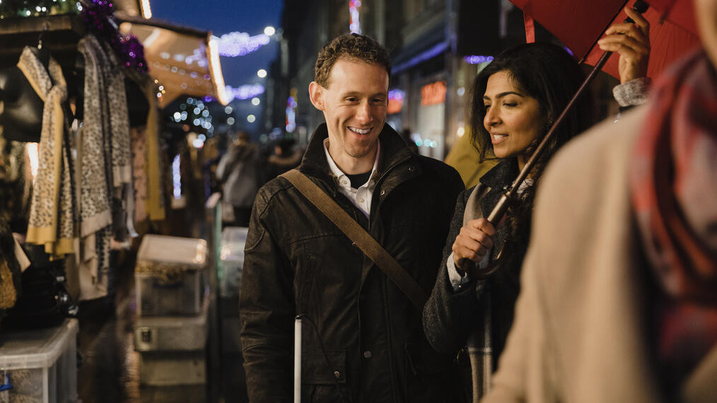 Visually impaired man walking alongside woman through market