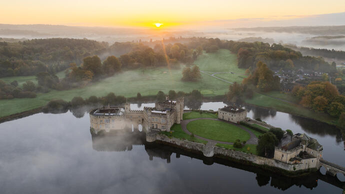 Vista aerea di un castello circondato da un fossato, un fiume e alberi, avvolto dalla nebbia all'alba