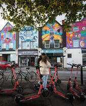 Two people standing by rental scooters in front of a row of houses painted on with colourful murals