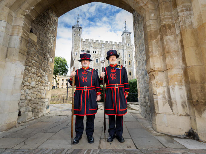 Two people in traditional Beefeater uniforms stand at a stone gate with the Tower of London in the background under a blue sky.