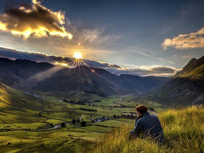 Man sitting in long grass on the side of a fell watching the sunset