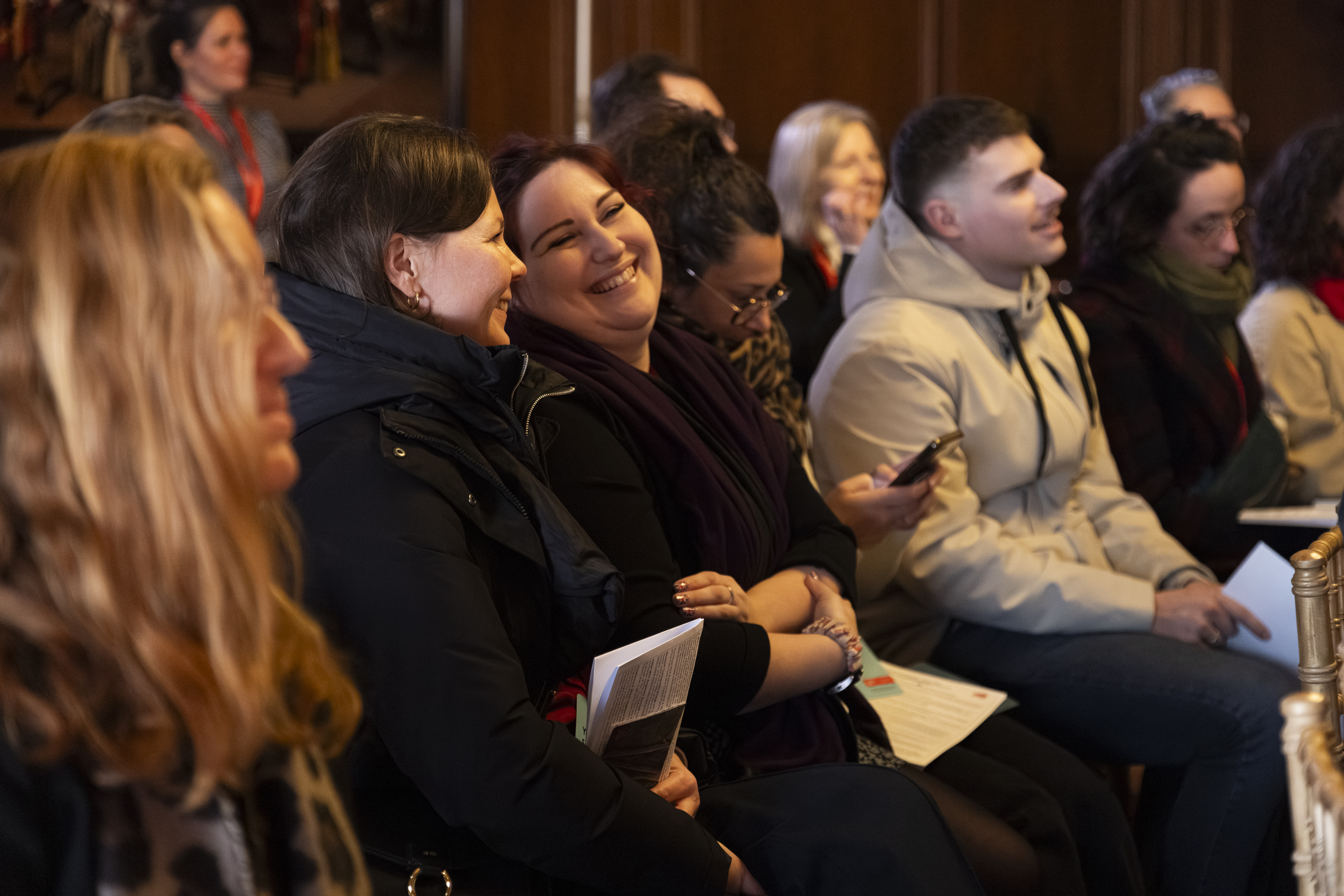 A group of people sitting indoors at an event, some smiling and laughing, with papers and phones in their hands.