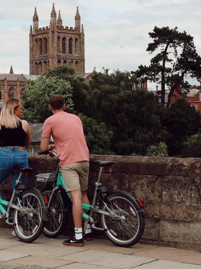 Deux personnes debout avec des vélos sur un pont en pierre, regardant une cathédrale historique entourée d'arbres et de bâtiments.