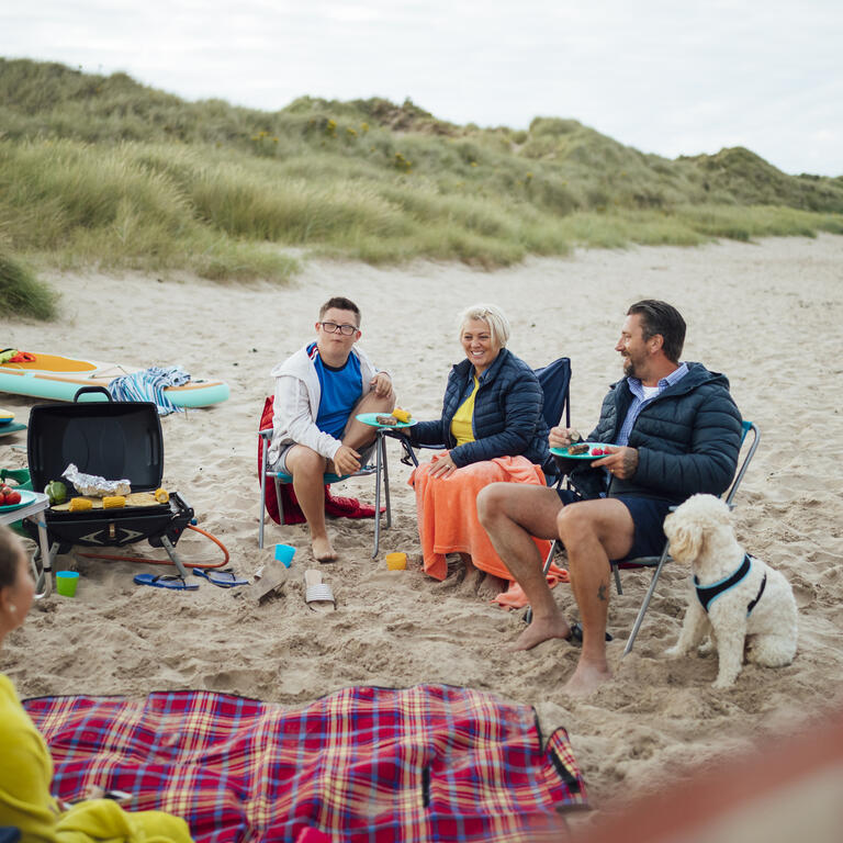 Una familia y su perro sentados junto a una manta de picnic en la playa.