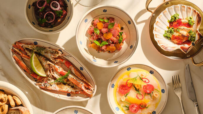 A variety of seafood and fresh dishes served on a marble table with bread and wine, presented in bright natural light.