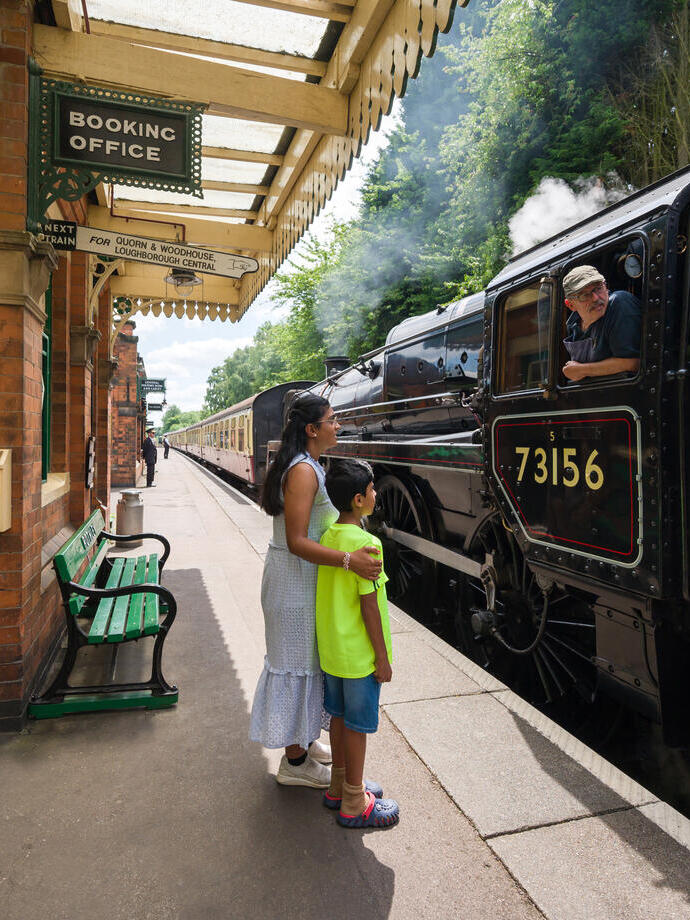 A woman and her son stand on a railway platform by a steam train