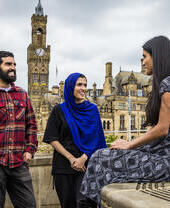 Two women and a man stand and talk in front of a town view