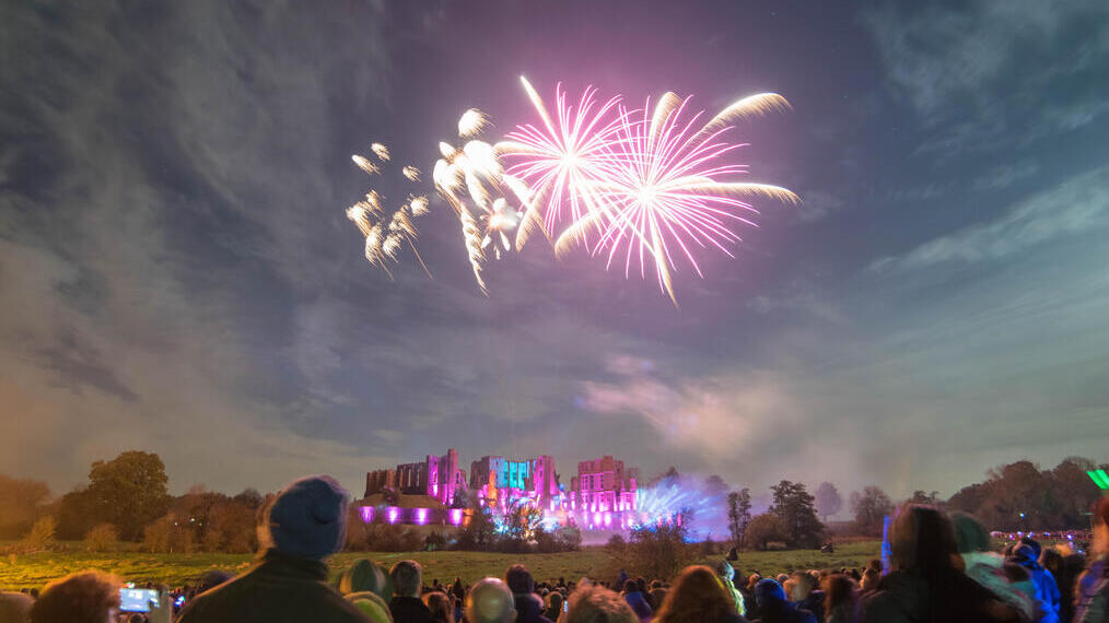 Fireworks display over a ruined castle watched by a crowd of people in the evening
