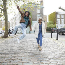 Happy young woman jumping while holding girlfriend's hand walking on a footpath in a street