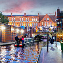 Riverboat cruising down a canal at dusk, with people walking down a parallel path.