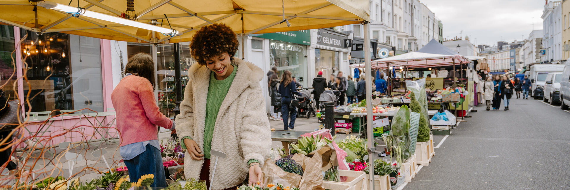 Outdoor flower market scene on a city street with people shopping at stalls under yellow tents, surrounded by historic buildings.