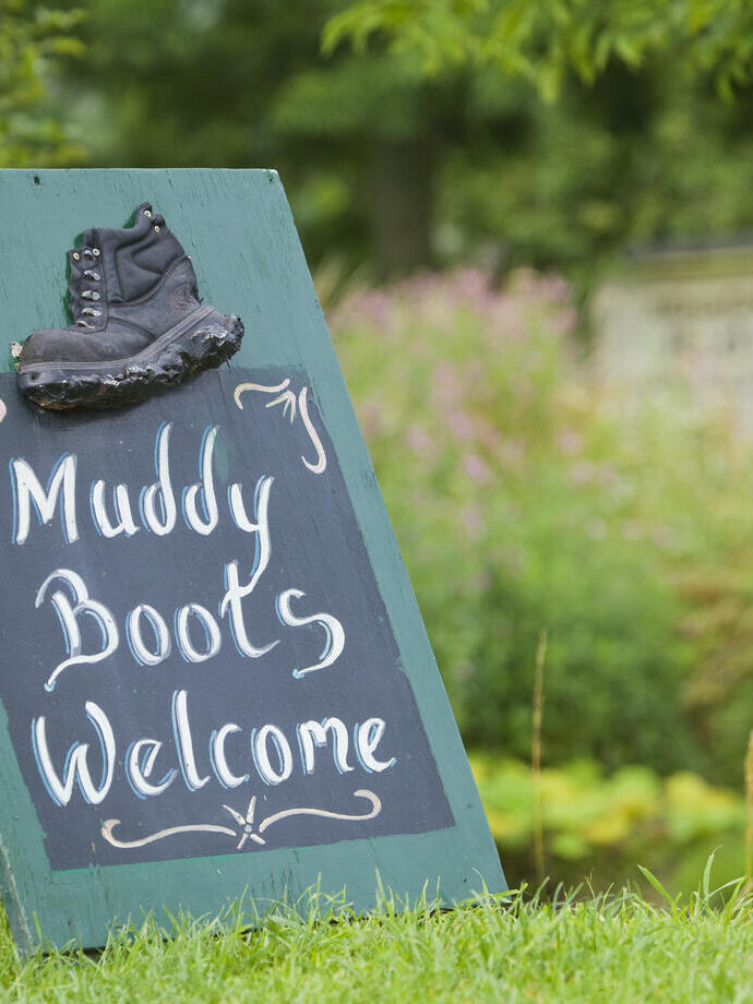 Signage on grass saying 'Muddy Boots Welcome' with plants