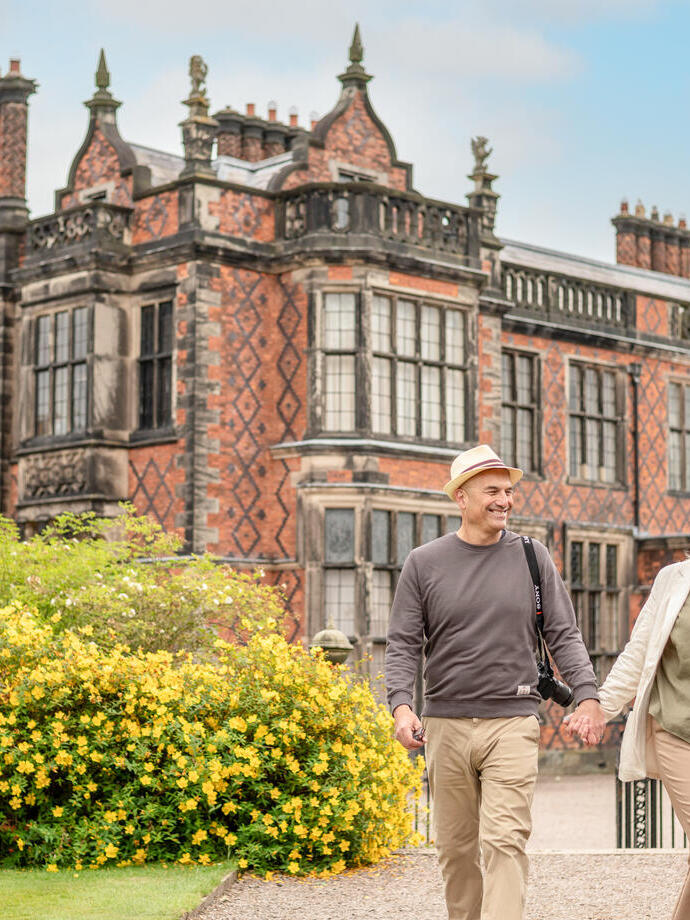 Un homme et une femme marchent côte à côte devant un manoir historique en briques rouges et sourient.