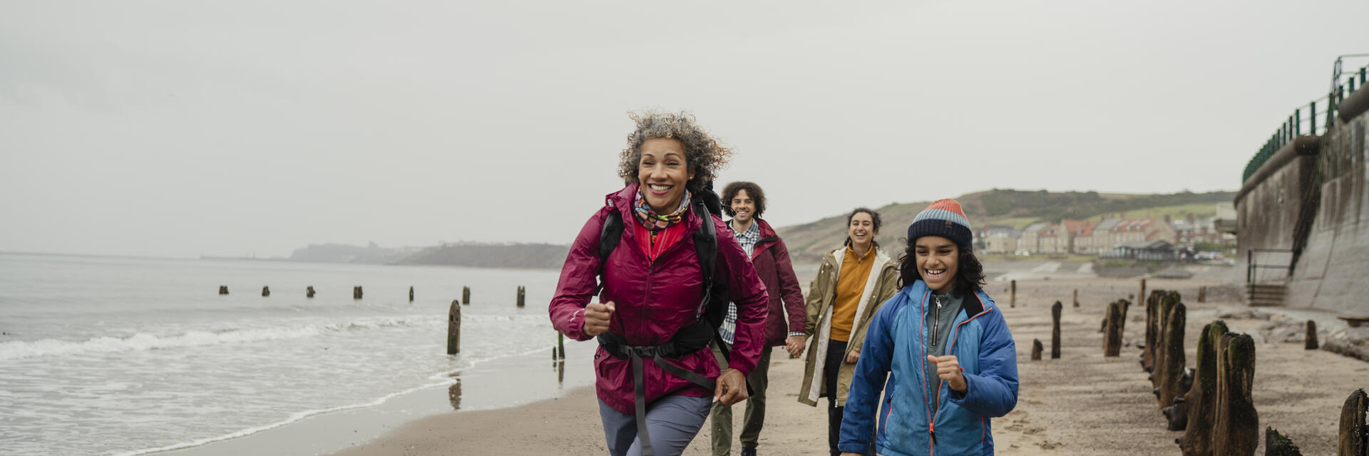 Group of people hiking and smiling along a sandy beach with wooden groynes, some houses, cliffs, and sea in the background on a cloudy day.