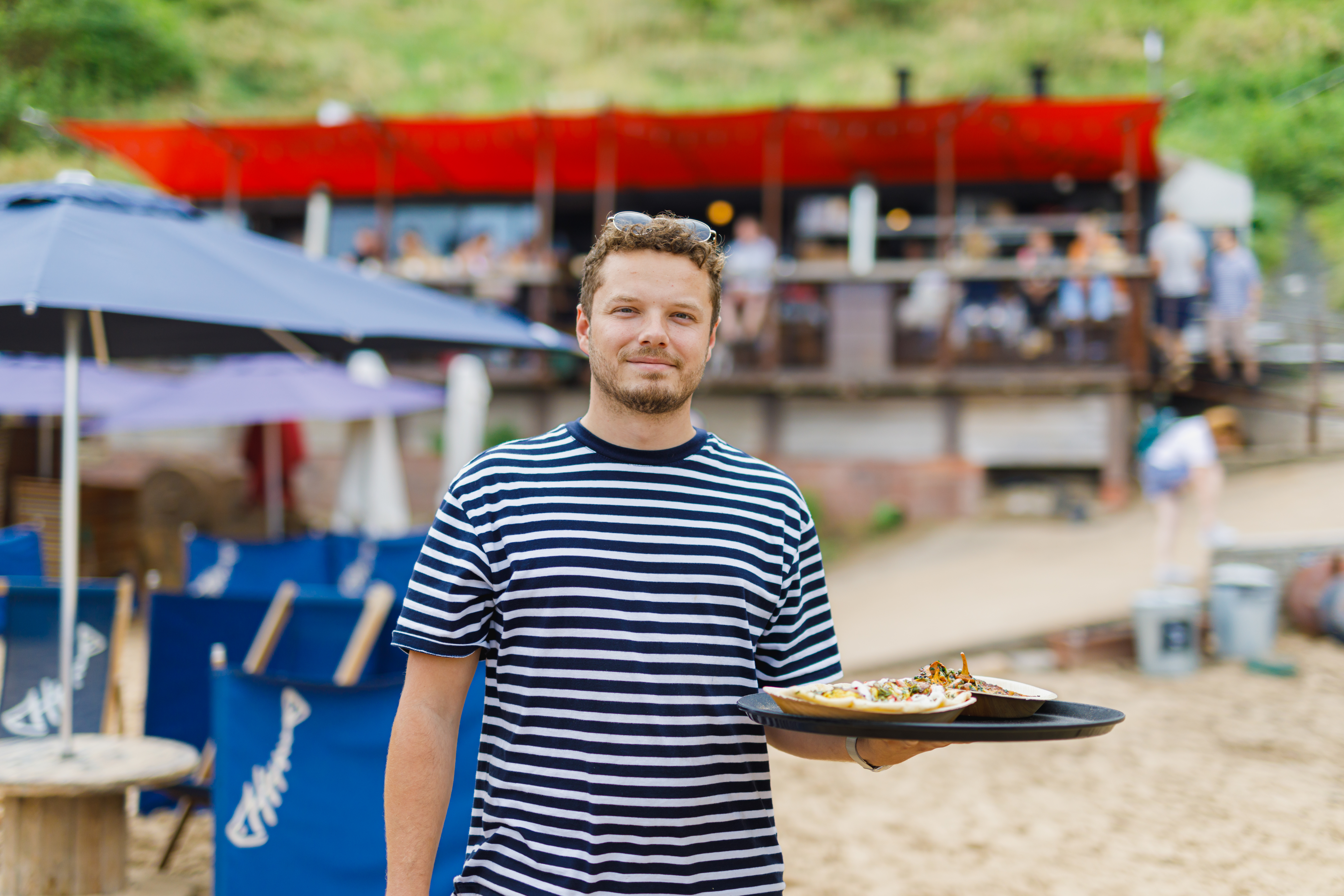 Hombre con camiseta de rayas lleva una bandeja de comida en la playa con restaurante al fondo.