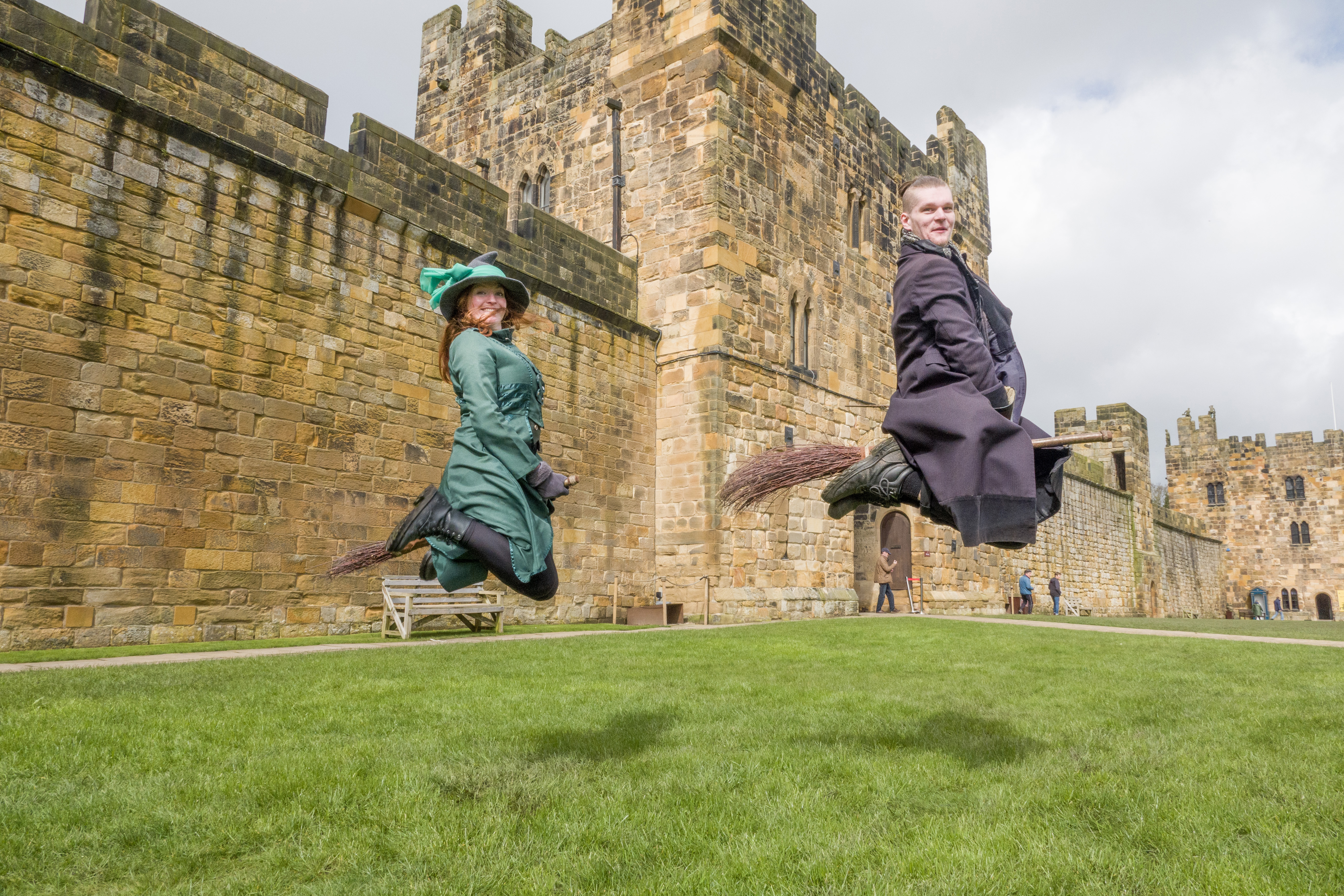 Two wizarding professors demonstrating flying on a broom outside a large castle.