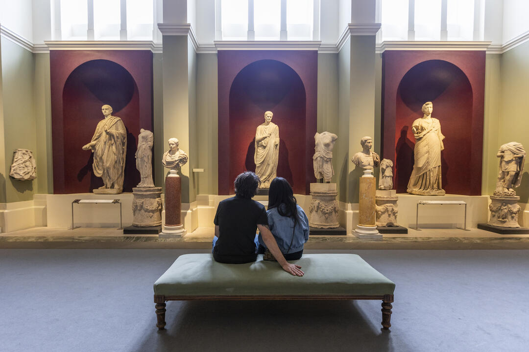 Two people sit on a bench in an art museum, viewing classical marble statues displayed in an elegant, well-lit gallery.