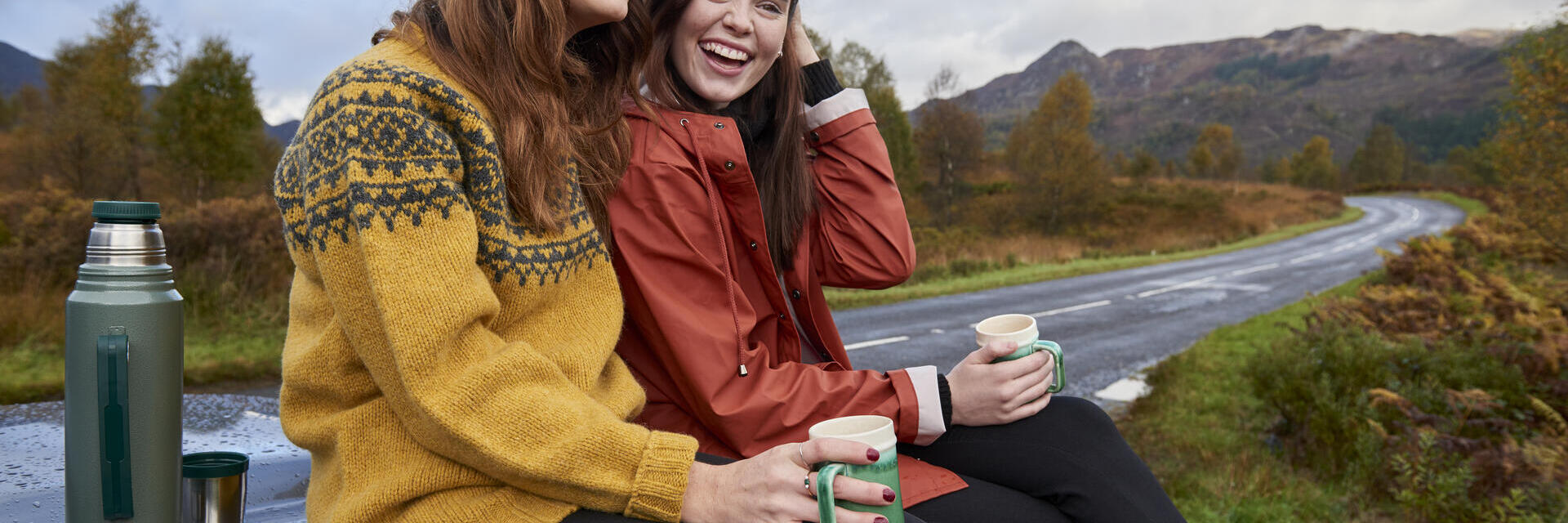 Zwei Personen sitzen auf einer Motorhaube, trinken aus Tassen in ländlicher Umgebung, mit Thermoskanne daneben und einer kurvigen Straße im Hintergrund.