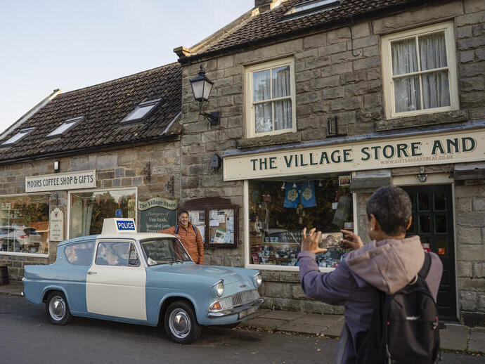 Street view of two people and a vintage police car outside a stone village store and coffee shop, with gift shop signs.