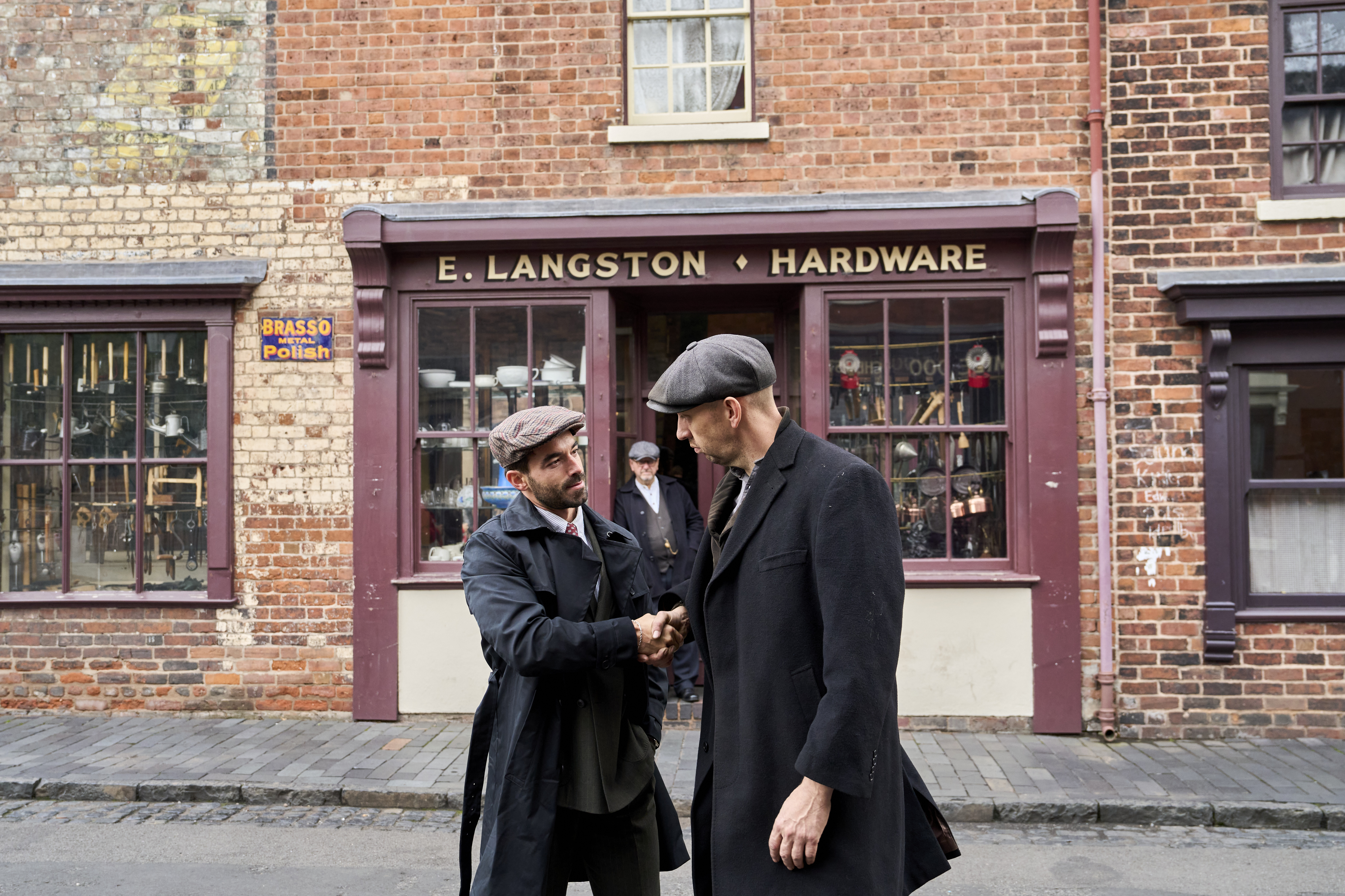 Two men in vintage attire shake hands outside a brick hardware store named E. Langston. Historical street scene with older shopfronts.