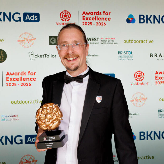 Person in formal attire holding a trophy at an awards event with branded backdrop for Awards for Excellence 2025-2026.