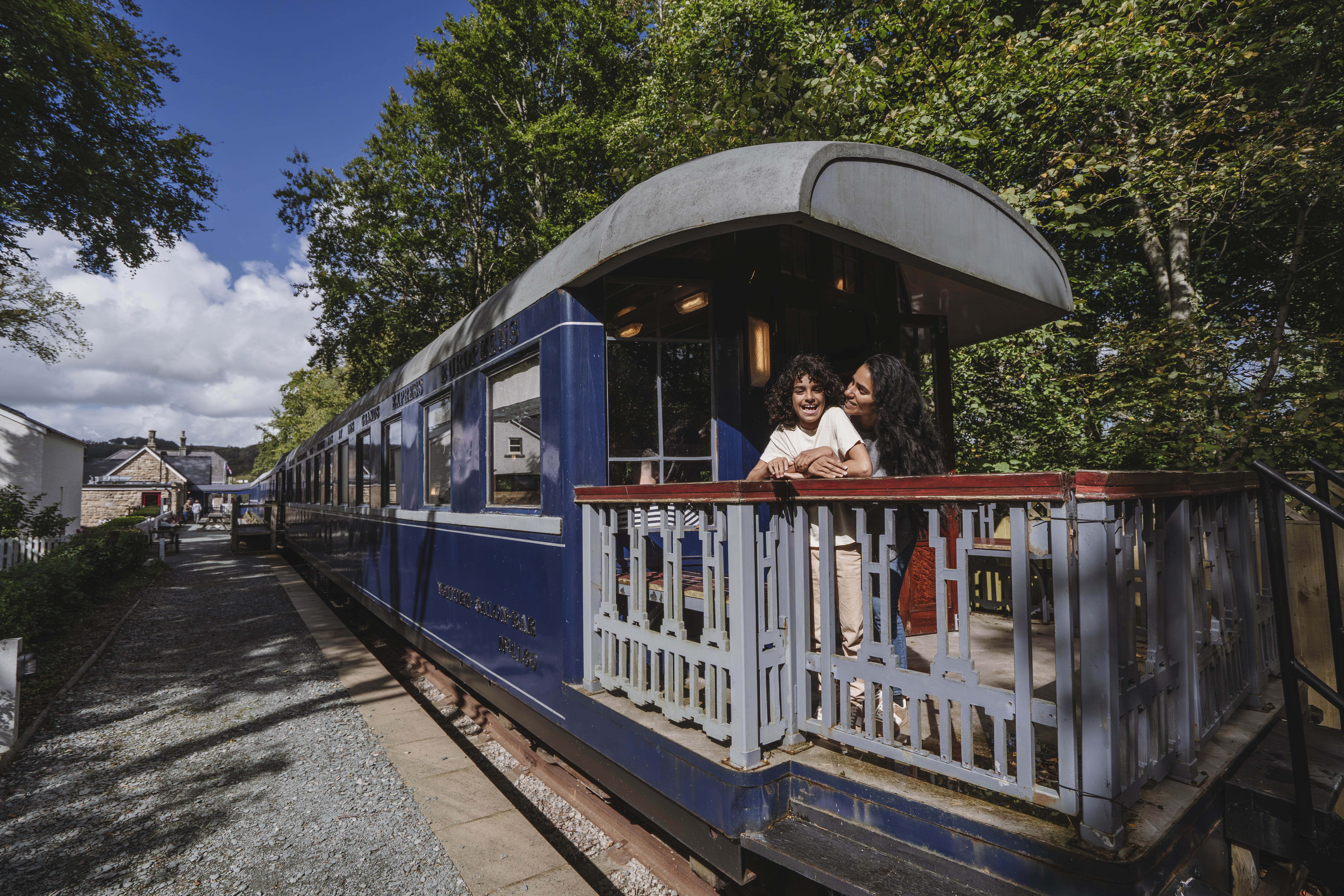 Historic blue train carriage at a station with trees around, people standing on the train's outdoor platform, and a house in the background on a sunny day.