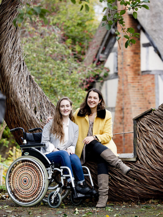 Two women, one in a wheelchair and one sitting on a large willow sculpture in a cottage garden