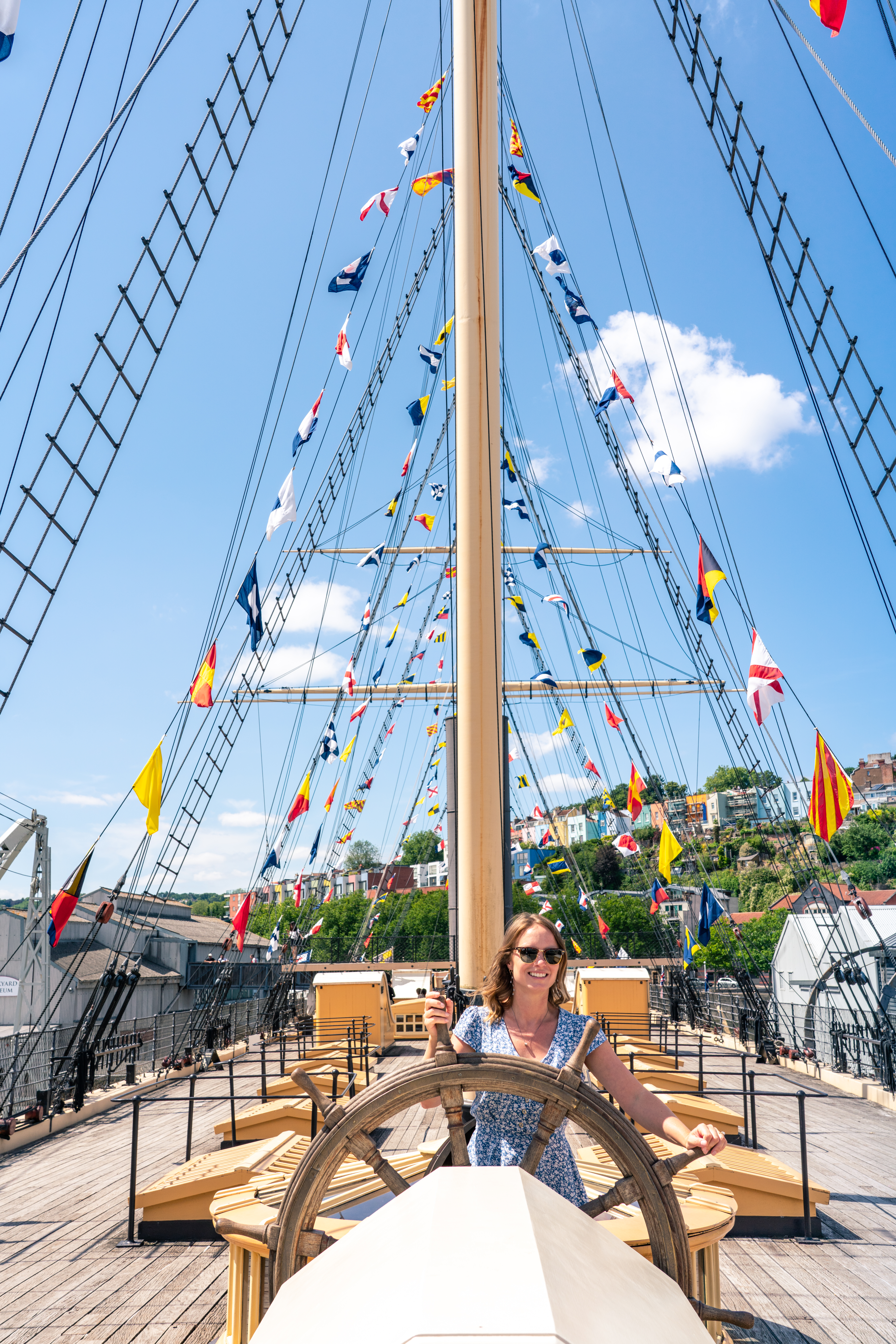 Smiling young woman in sunglasses at the wheel of a ship docked on land with flags on guide ropes against blue sky
