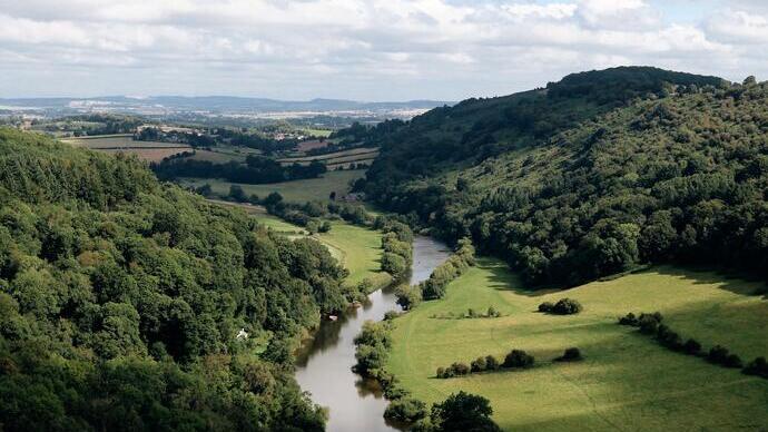 A river winding through a lush green valley with forested hills on both sides and a cloudy sky above.