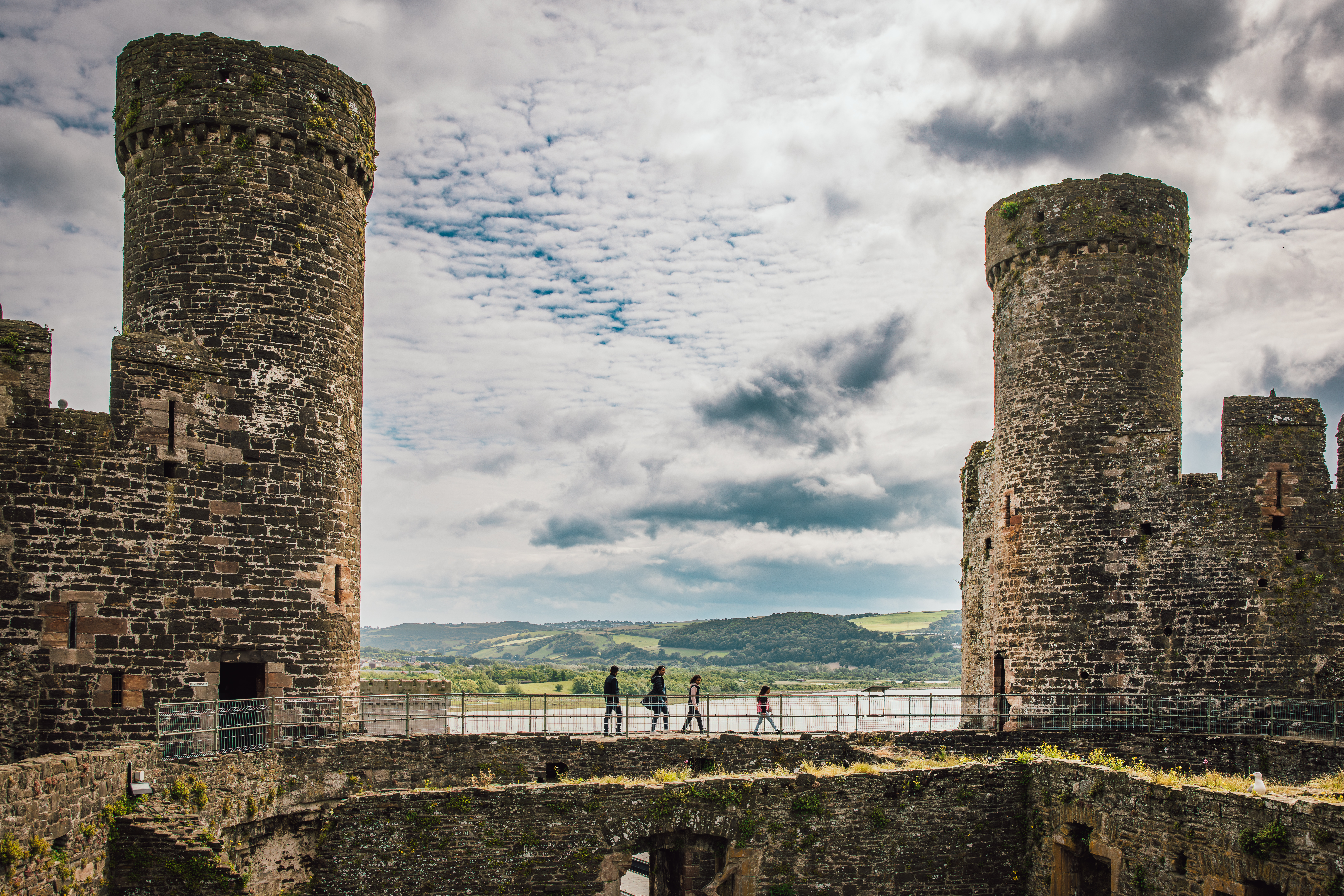 Stone castle ruins with two large round towers, people walking on a wall, hills and water visible in the background under a cloudy sky.