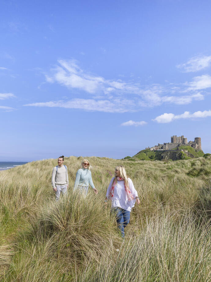 A group of people walk among tall grasses in dunes with a castle in the background