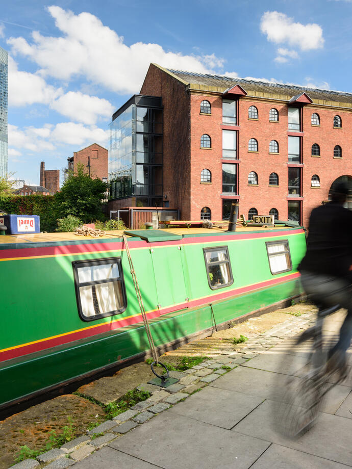 Canal boat moored by waterside, red brick warehouse, modern skyscraper, cyclists passing, sunny urban scene.