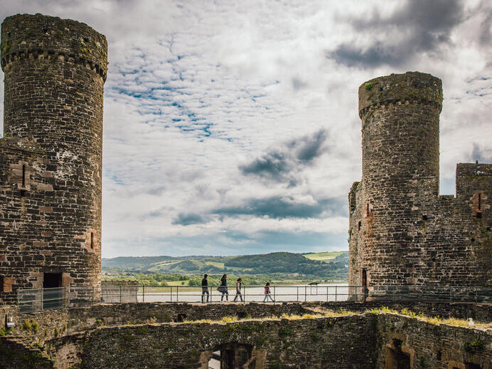Stone castle ruins with two large round towers, people walking on a wall, hills and water visible in the background under a cloudy sky.