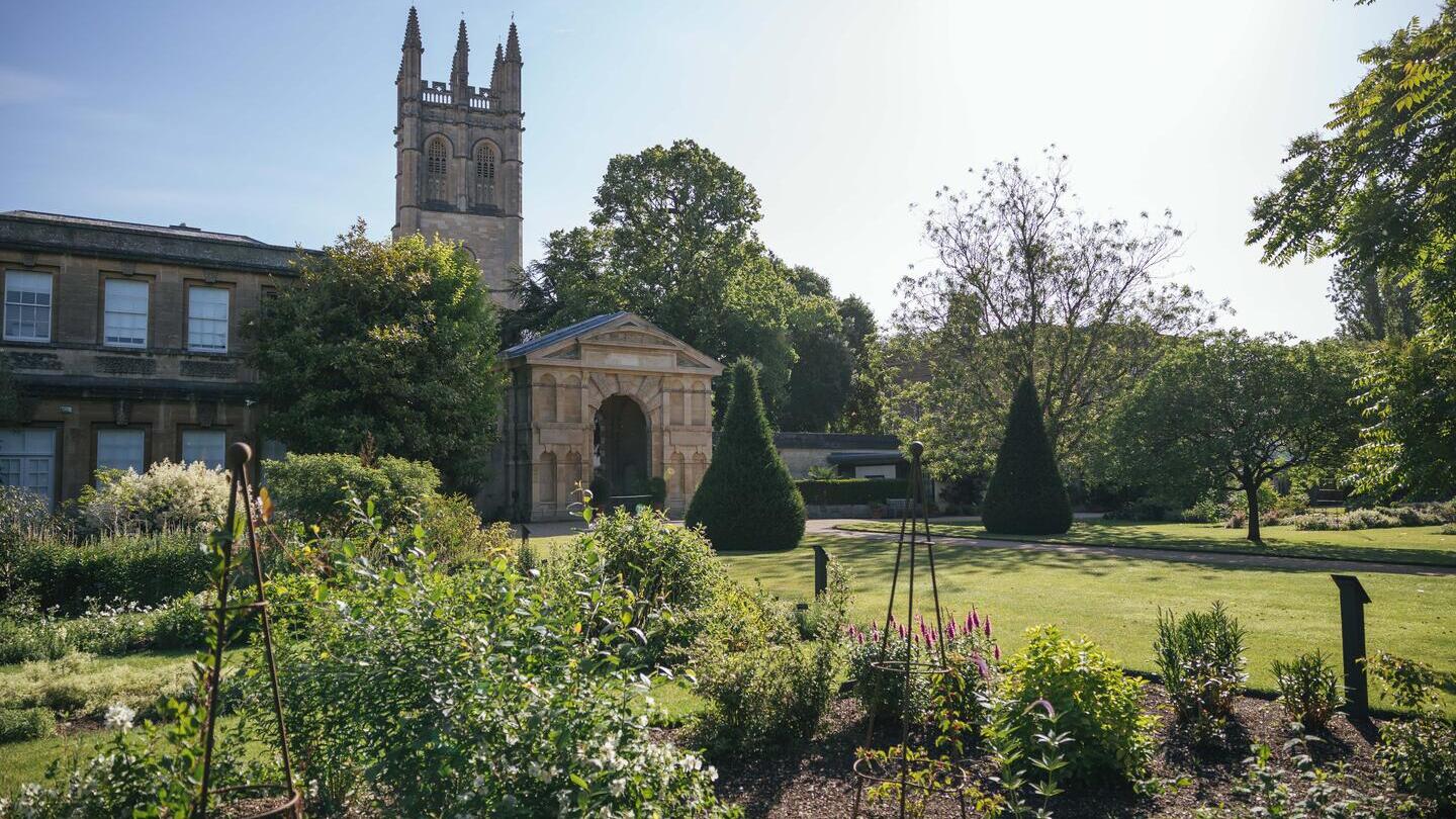 Flower bed in front of a georgian arch and building of Oxford Botanic Garden with church tower behind against blue sky