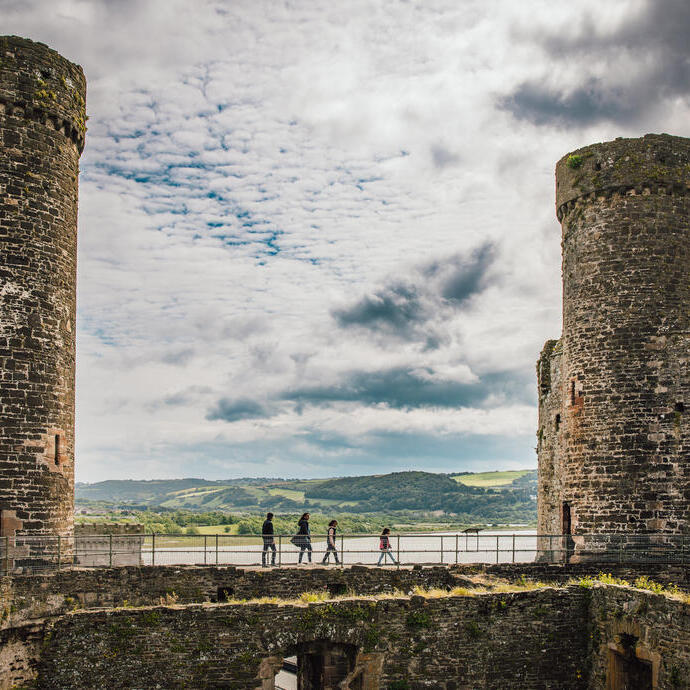 Stone castle ruins with two large round towers, people walking on a wall, hills and water visible in the background under a cloudy sky.