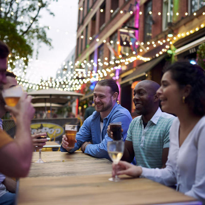 Amigos tomando una copa sentados en una mesa exterior de un bar con guirnaldas de luces colgando detrás