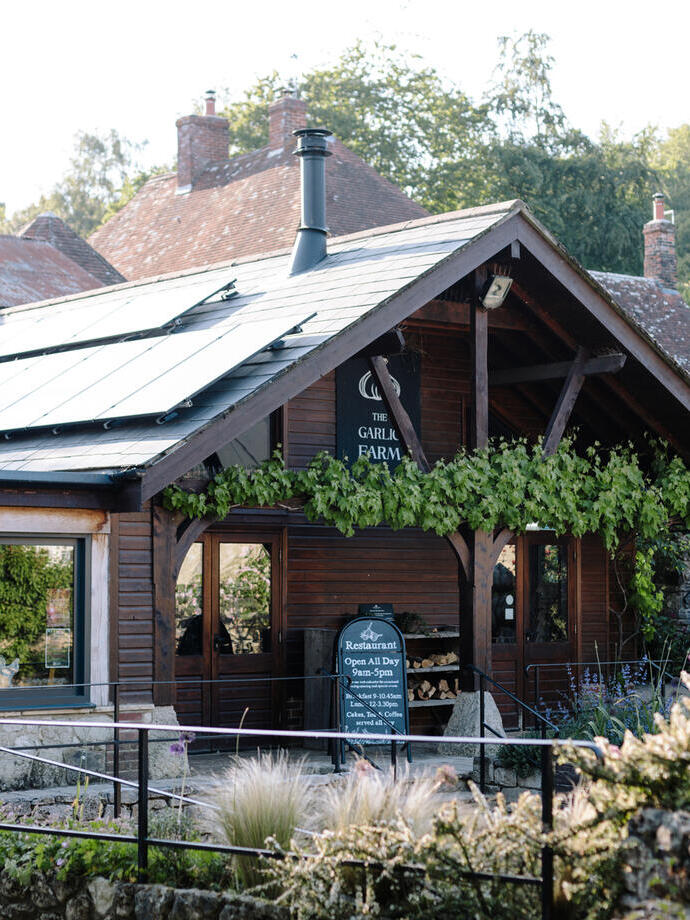 Charming wooden restaurant building with a vine-wrapped porch, surrounded by lush gardens and greenery in a rural setting.