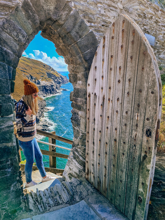 A woman stands at the top steps of a doorway with coastline view beyond