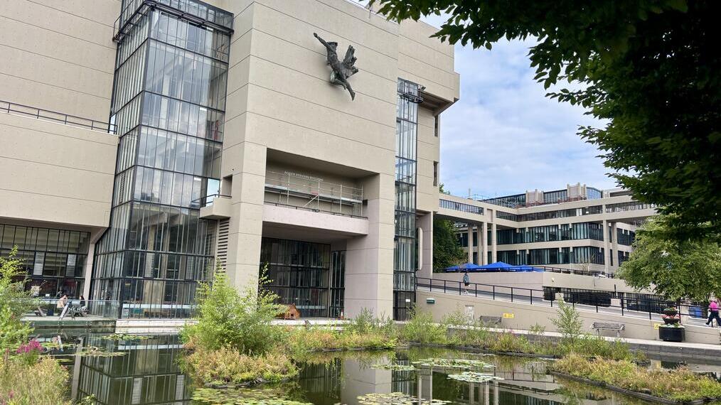 Modern concrete building with glass sections, a sculpture on the facade, pond with lily pads in foreground, greenery, and people nearby.